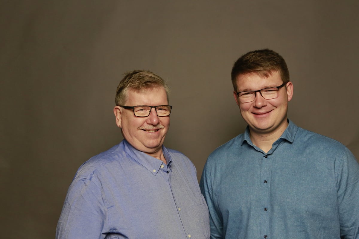 Two men wearing glasses and button-up shirts stand together smiling against a neutral gray background