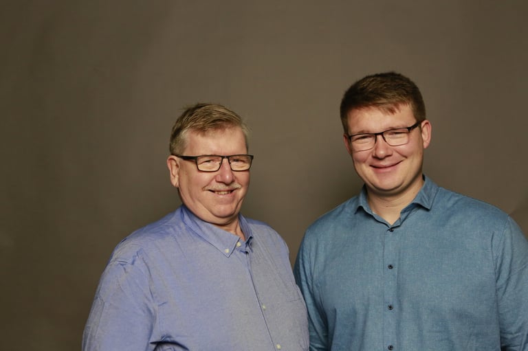 Two men wearing glasses and button-up shirts stand together smiling against a neutral gray background