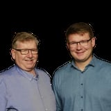 Two men wearing glasses and casual button-up shirts smiling at the camera against a black background
