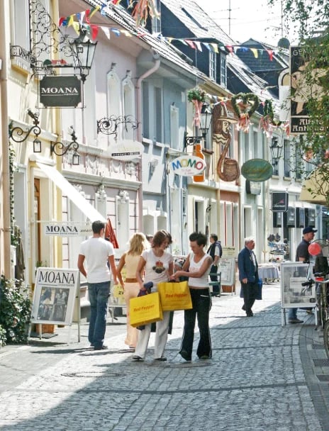 Charming European shopping street with white buildings, boutique storefronts, and shoppers with yellow bags strolling on a sunny day