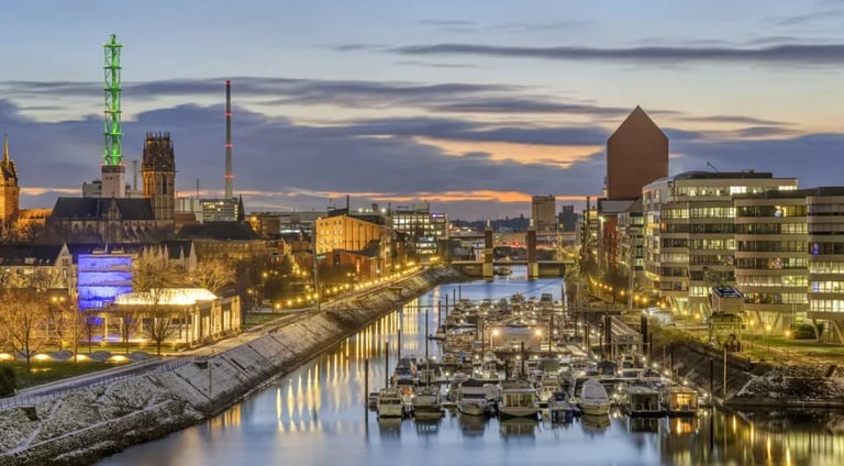 Twilight view of a waterfront city with illuminated historic buildings, moored sailboats, and a church spire reflecting in calm water